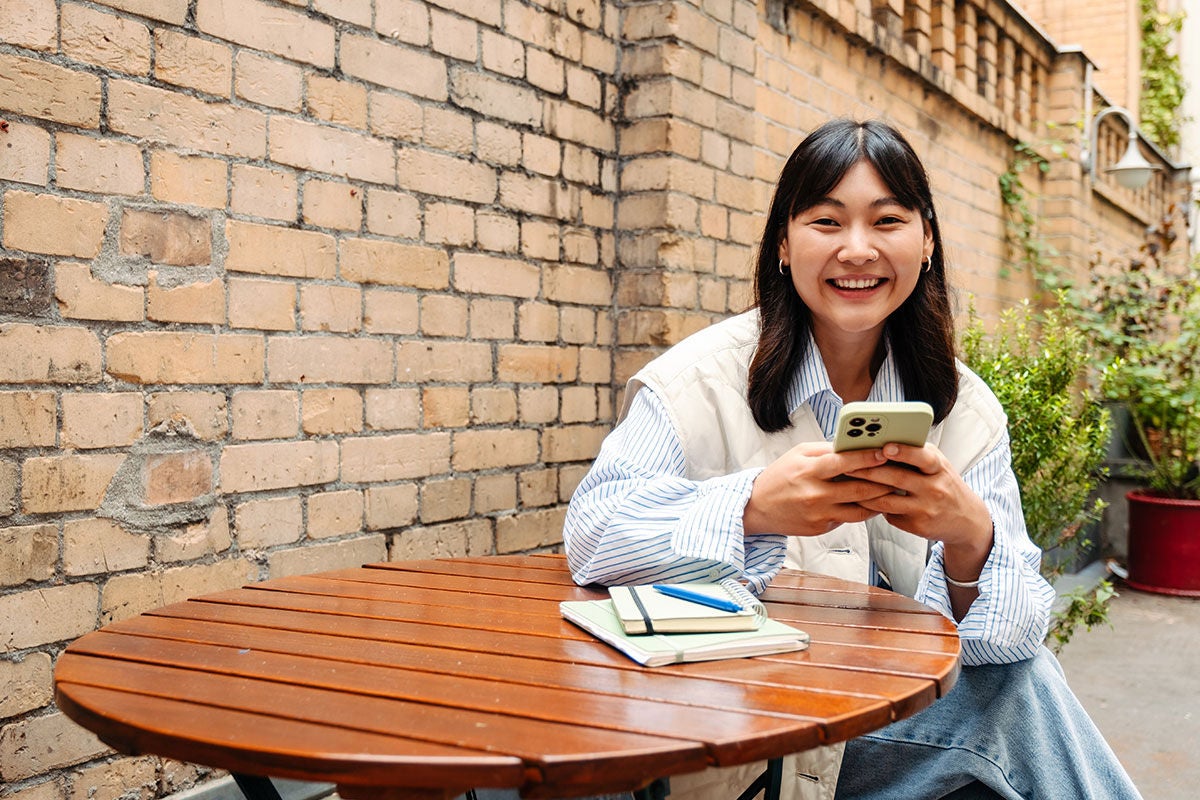 A woman sits at a table holding a mobile phone. She is outside and smiling at the camera.