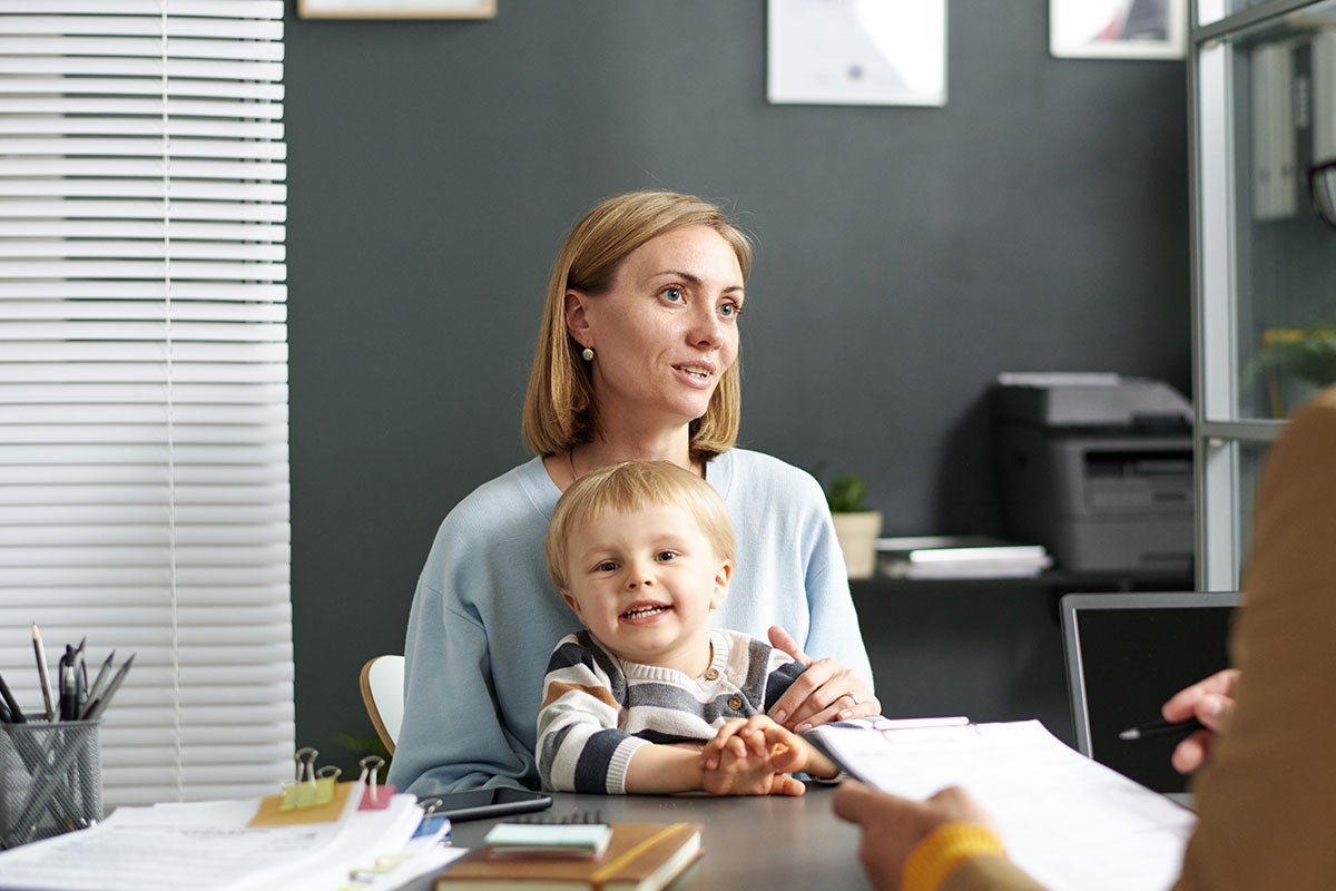 A woman sits at a desk in conversation with a person whose back is to camera. On the woman's lap is a smiling child. The desk is covered with various papers and office materials.