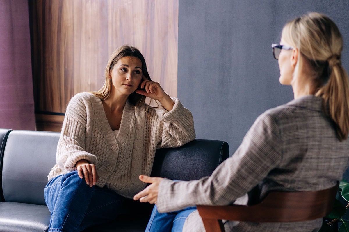 A woman sits on a lounge. She is intently listening to another woman who sits opposite her with her back to the camera. 